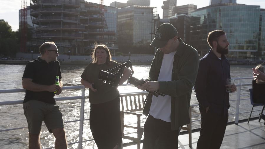 A videographer films with a gimbal-mounted camera while guests chat on deck in the evening sun